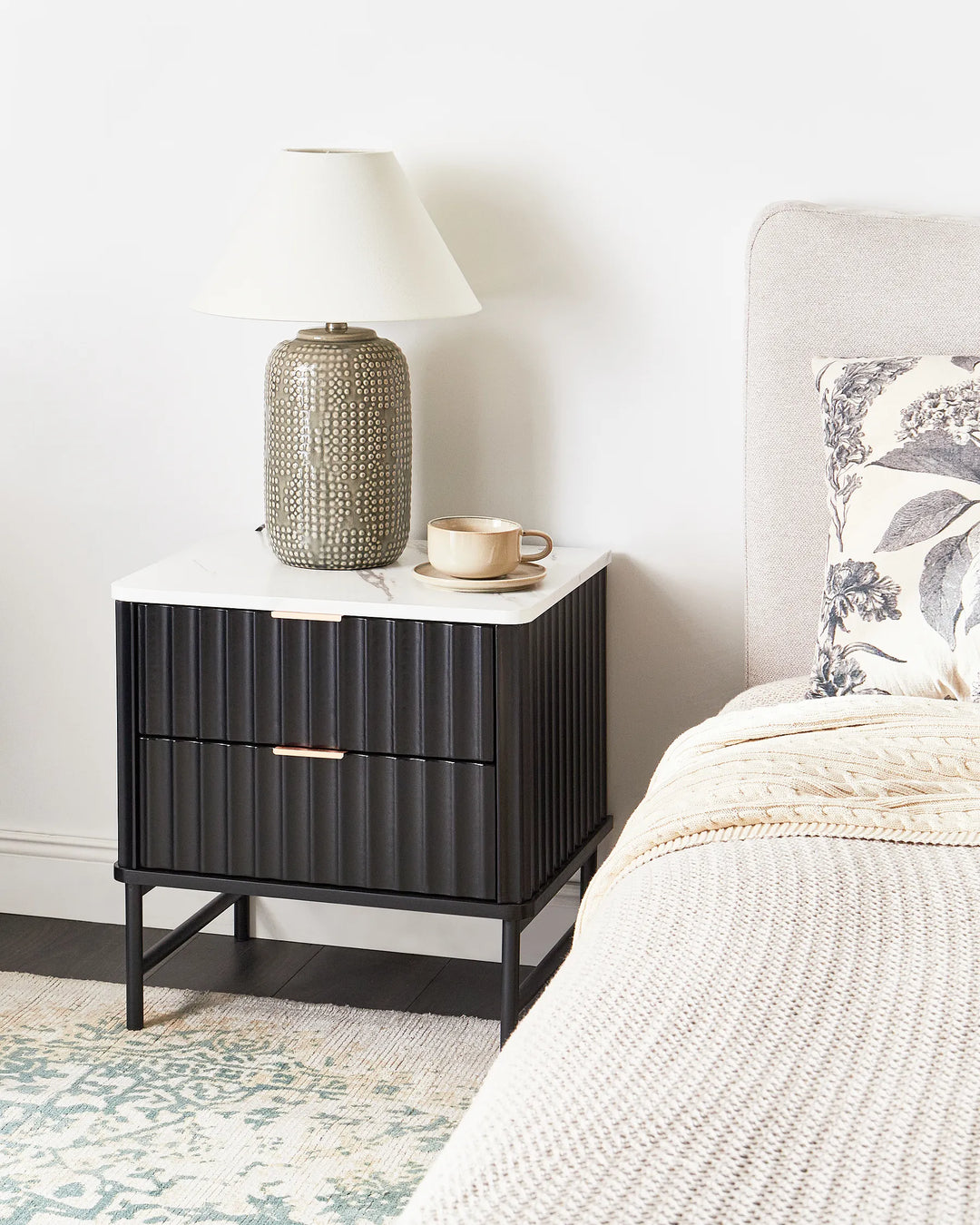 Black nightstand with white top next to a bed with a floral pillow