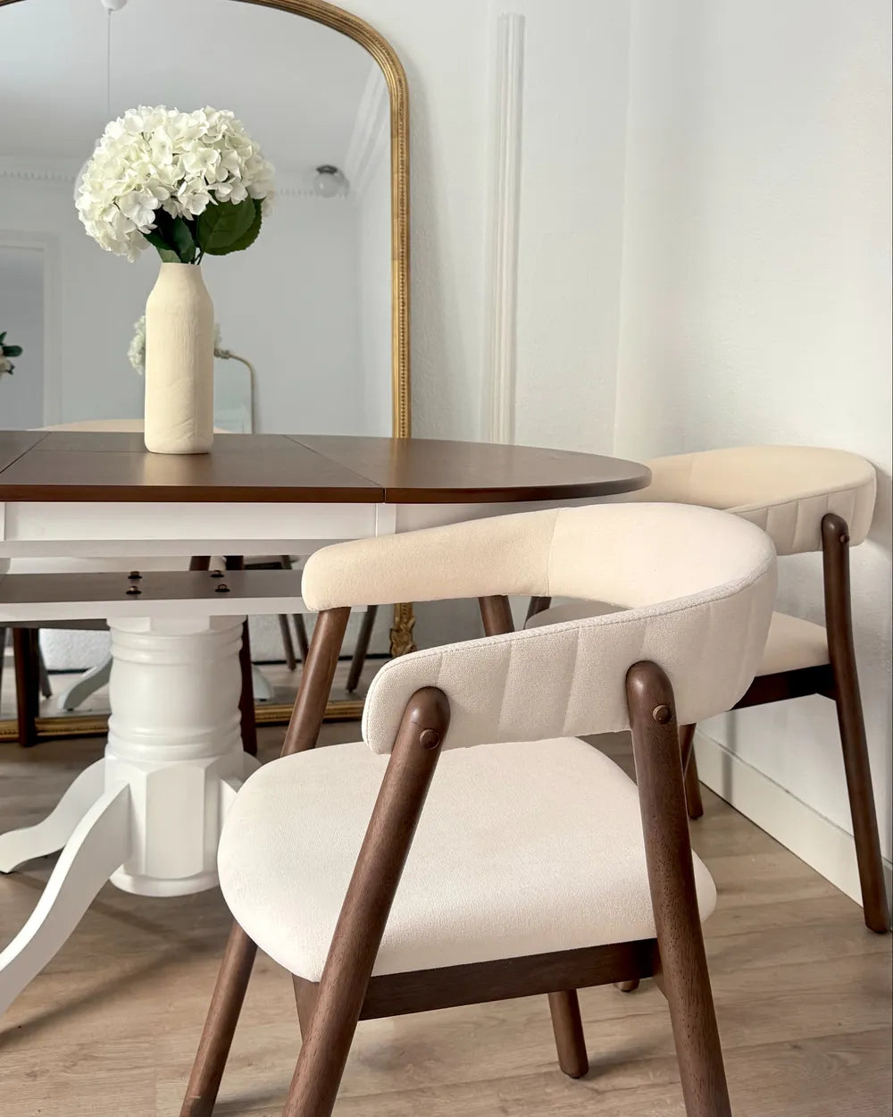 A dining room scene featuring two modern armchairs with cream upholstered seats and dark wood frames, positioned around a white pedestal table with a gold-framed mirror and a vase of white flowers in the background.