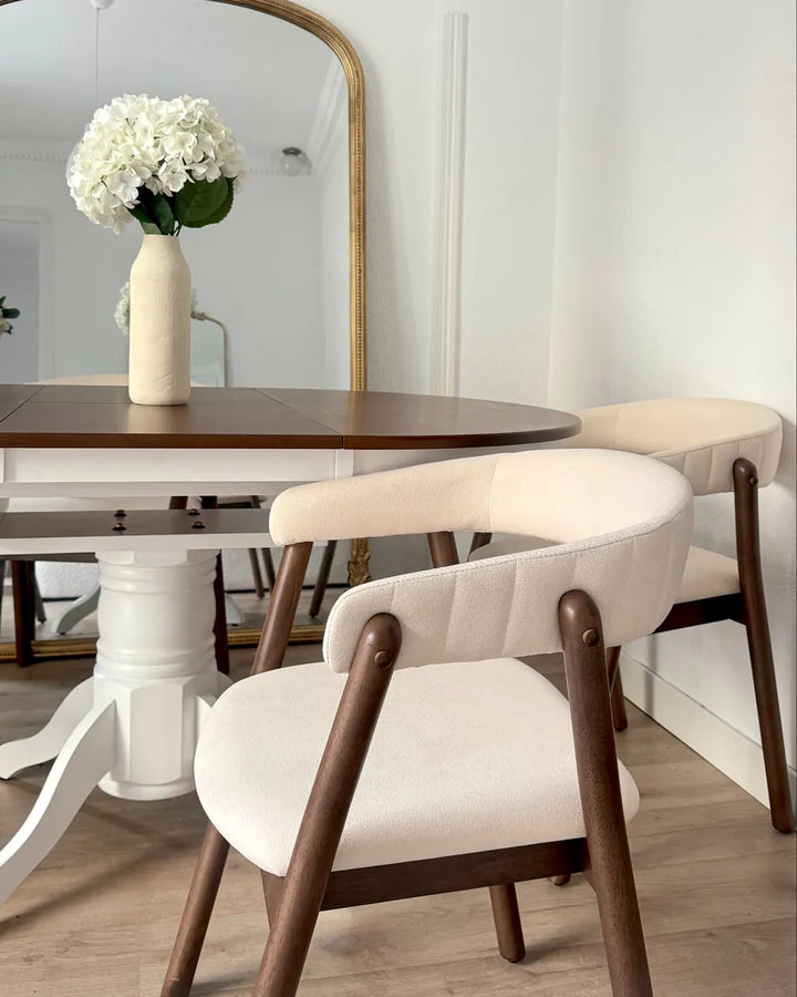 A dining room scene featuring two modern armchairs with cream upholstered seats and dark wood frames, positioned around a white pedestal table with a gold-framed mirror and a vase of white flowers in the background.