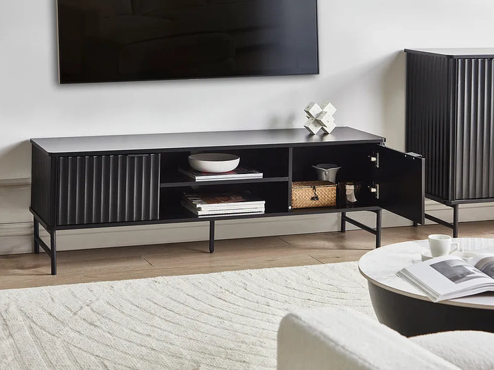 A modern black TV console cabinet with a ribbed front, open center shelf, and open side cabinet door, styled in a living room setting with a TV, rug, and coffee table.