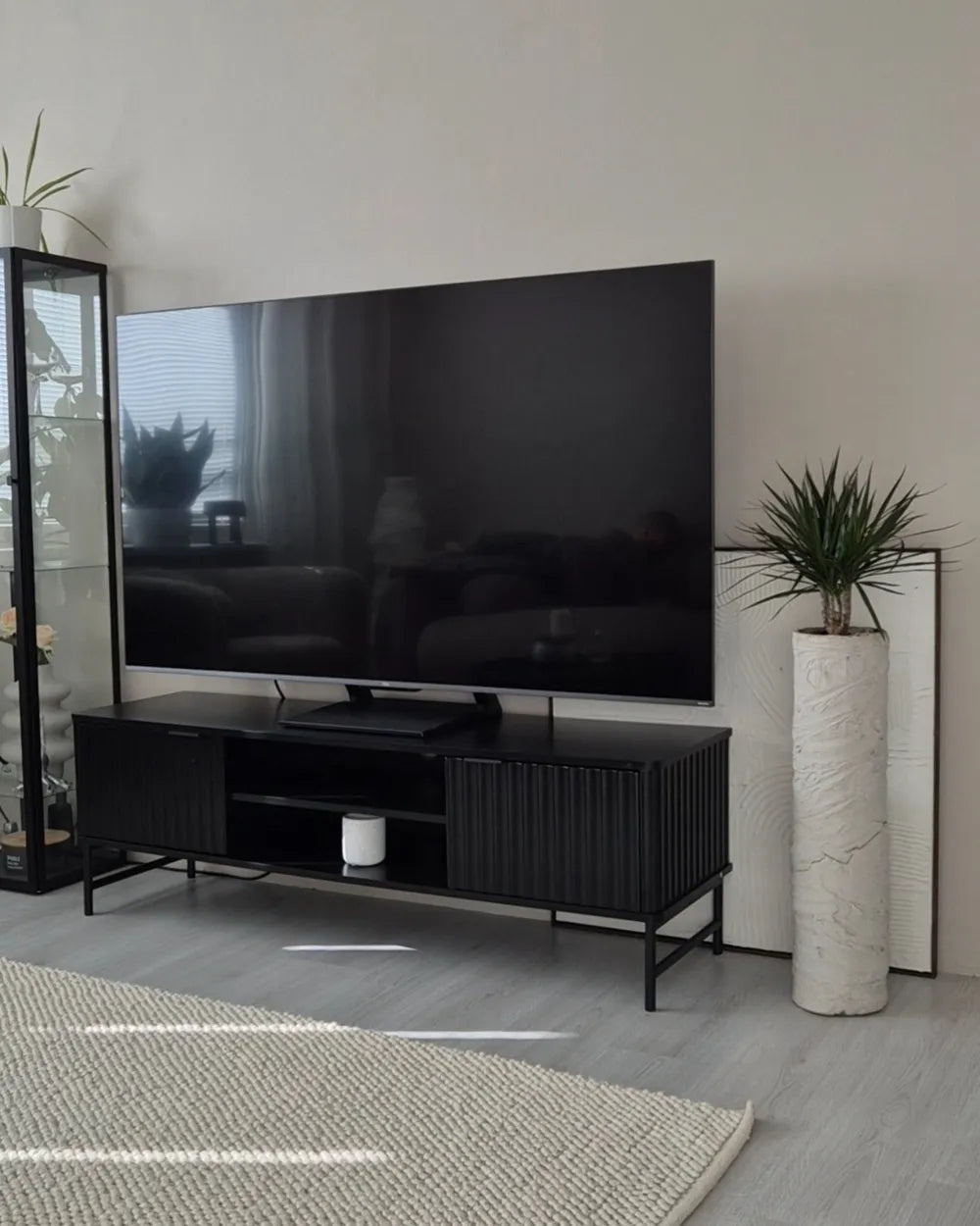 A modern black TV stand with a flat-screen television on top, styled in a minimalist living room with a light gray wall, gray wood floor, a white marble pillar, and a woven rug.
