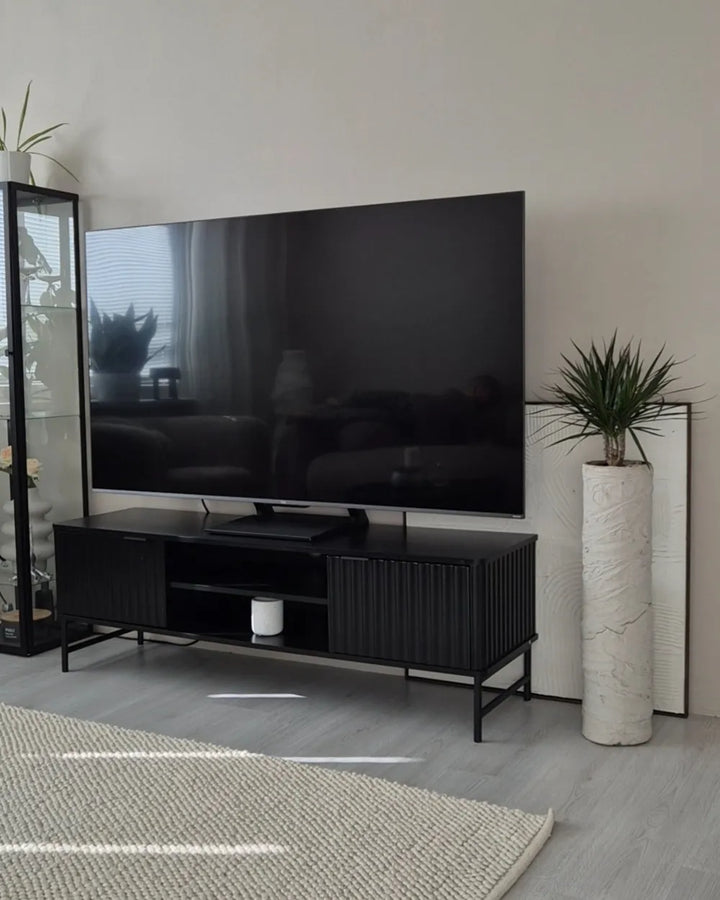 A modern black TV stand with a flat-screen television on top, styled in a minimalist living room with a light gray wall, gray wood floor, a white marble pillar, and a woven rug.