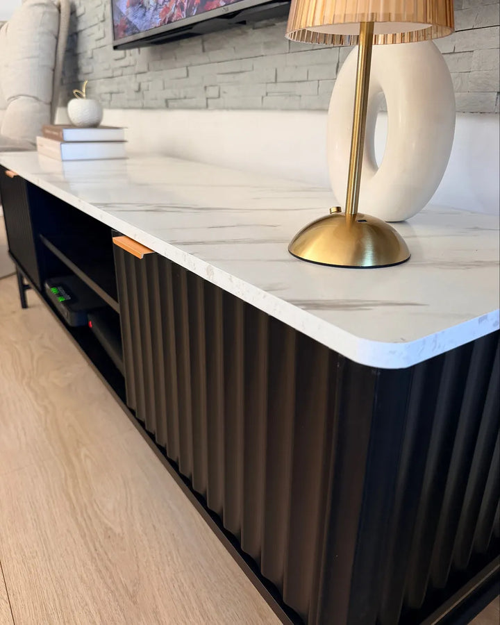 A modern TV stand or media console with a white marble-look top, black ribbed side panels, open shelving, and a brass lamp placed on top in a styled interior setting.