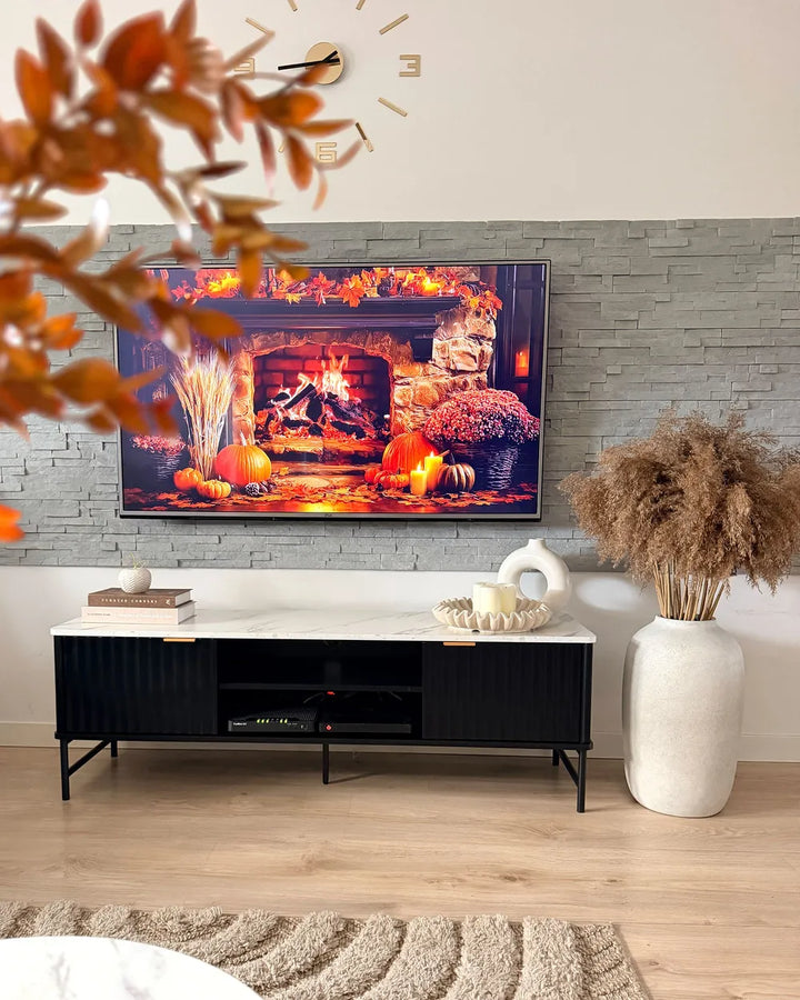 A modern living room setup featuring a black media console with a white marble top, displaying a television set showing a cozy fireplace scene. The console is styled with books, a small sculpture, and a large white vase with dried pampas grass on a light wood floor.