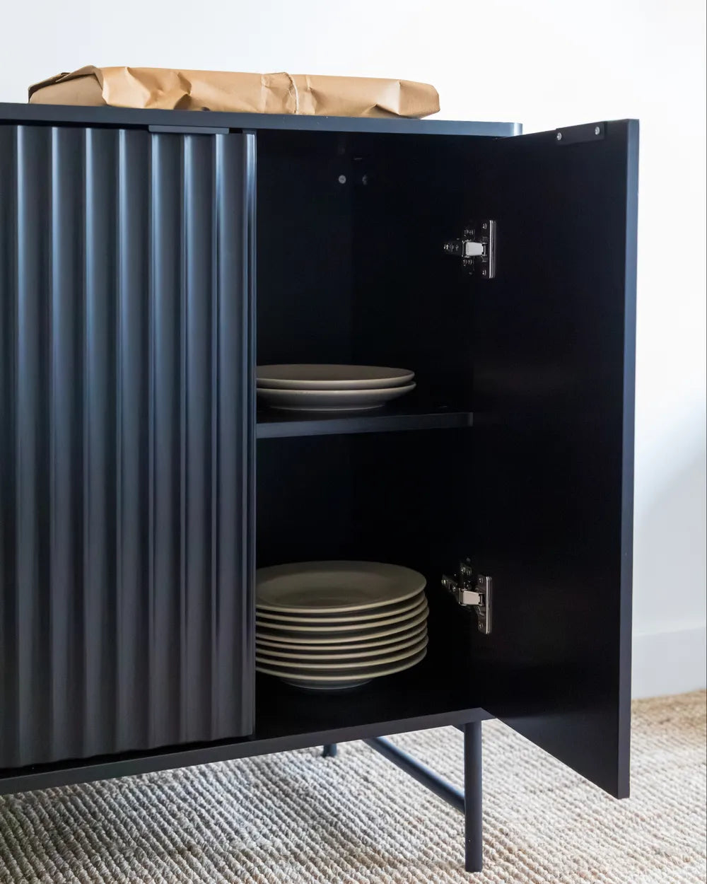 A modern black sideboard cabinet with a ribbed front door and open hinged side door, shown holding plates on a shelf. The cabinet stands on thin metal legs over a textured rug, with a brown paper bag on top against a white wall.