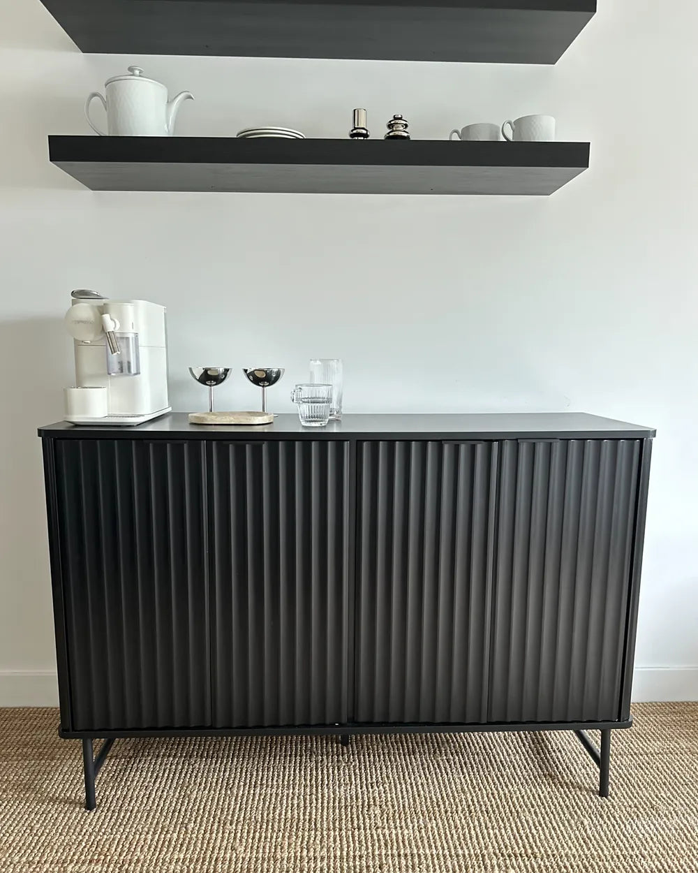 A modern black ribbed sideboard cabinet with tapered legs, styled in a minimalist room with floating shelves and home decor on top, set against a white wall and a woven rug.