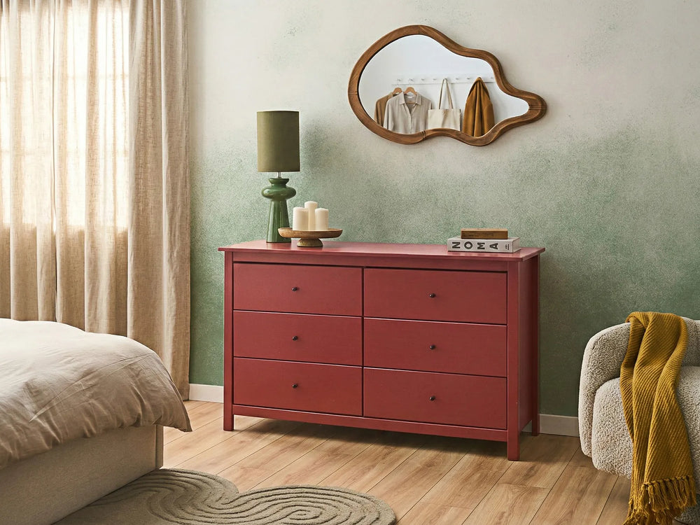 A six-drawer wooden dresser in a dusty rose pink finish, styled in a bedroom setting with a green lamp, white candles, a cloud-shaped mirror, a bed, and a cozy armchair.