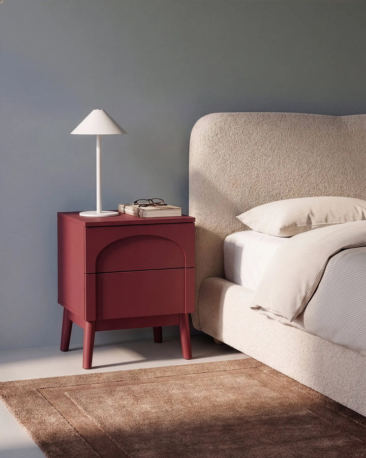 A modern bedroom scene featuring a dark red nightstand with a white table lamp and books, positioned next to a bed with a textured beige headboard and white bedding, set against a muted blue-gray wall.