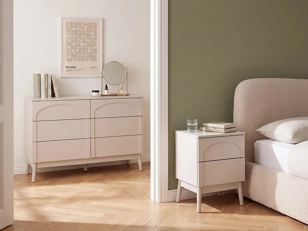 A light beige bedroom furniture set shown in a styled room, featuring a six-drawer dresser against a white wall and a matching two-drawer nightstand beside a bed with a sage green wall.
