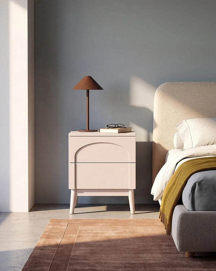 A modern two-drawer nightstand in a pale blush pink finish stands beside a bed in a minimalist bedroom. A dark brown table lamp and a small stack of books sit on top of the nightstand.