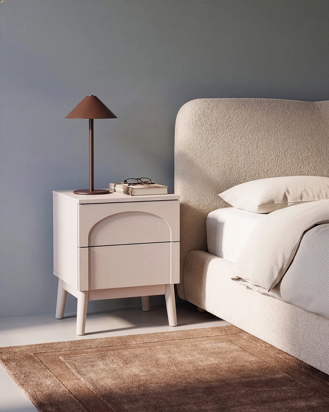 A modern cream-colored two-drawer nightstand with a curved drawer front, styled in a bedroom next to a bed with white linens and a brown table lamp on top.