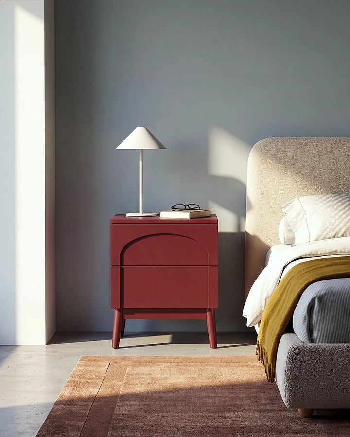 A modern bedroom scene featuring a dark red nightstand with a curved front detail, styled with a white table lamp and glasses. The nightstand is positioned next to a bed with white linens and a yellow throw blanket against a muted blue-gray wall.