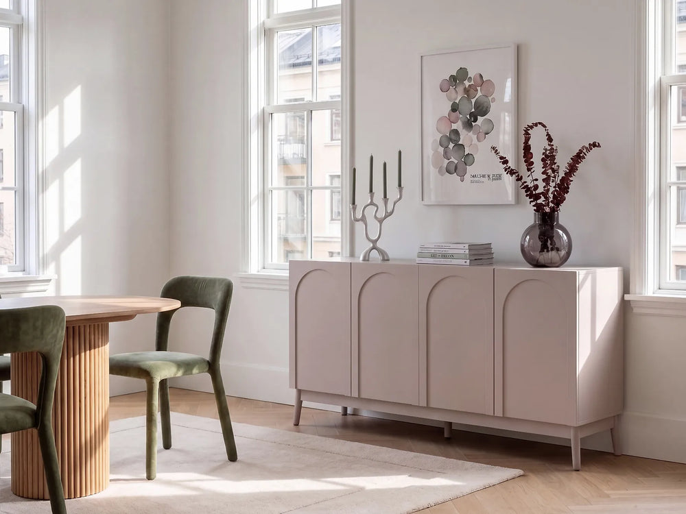 A light blush pink sideboard cabinet with arched panel doors in a bright, modern room. The cabinet is styled with a silver candelabra, a glass vase with dried branches, and a framed botanical print on the wall.