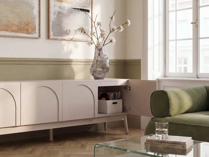 A modern cream-colored sideboard cabinet with arched panel doors and a glass vase of branches on top, shown in a styled living room next to a green sofa.