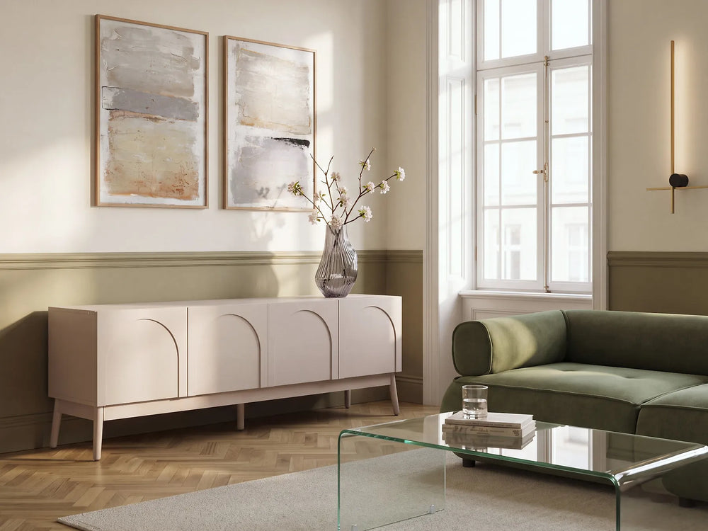 A modern living room scene featuring a pale blush pink sideboard cabinet with arched panel doors, styled with a glass vase of branches. The room has cream walls with green wainscoting, a green velvet sofa, and a glass-top coffee table.