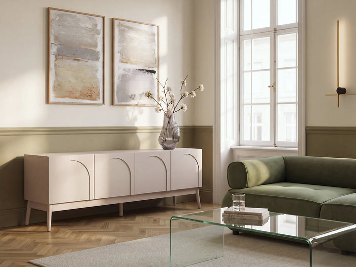 A modern living room scene featuring a pale blush pink sideboard cabinet with arched panel doors, styled with a glass vase of branches. The room has cream walls with green wainscoting, a green velvet sofa, and a glass-top coffee table.