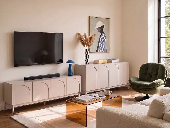 A modern living room featuring a light blush pink double cabinet TV stand and a matching sideboard with arched door details, styled with a wall-mounted TV, a green velvet armchair, and a translucent amber coffee table.
