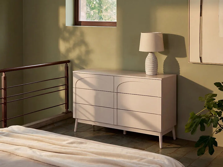 A modern light beige dresser with curved drawer fronts and tapered legs, styled in a bedroom next to a bed with cream bedding, a textured white table lamp, and a green plant against a sage green wall.