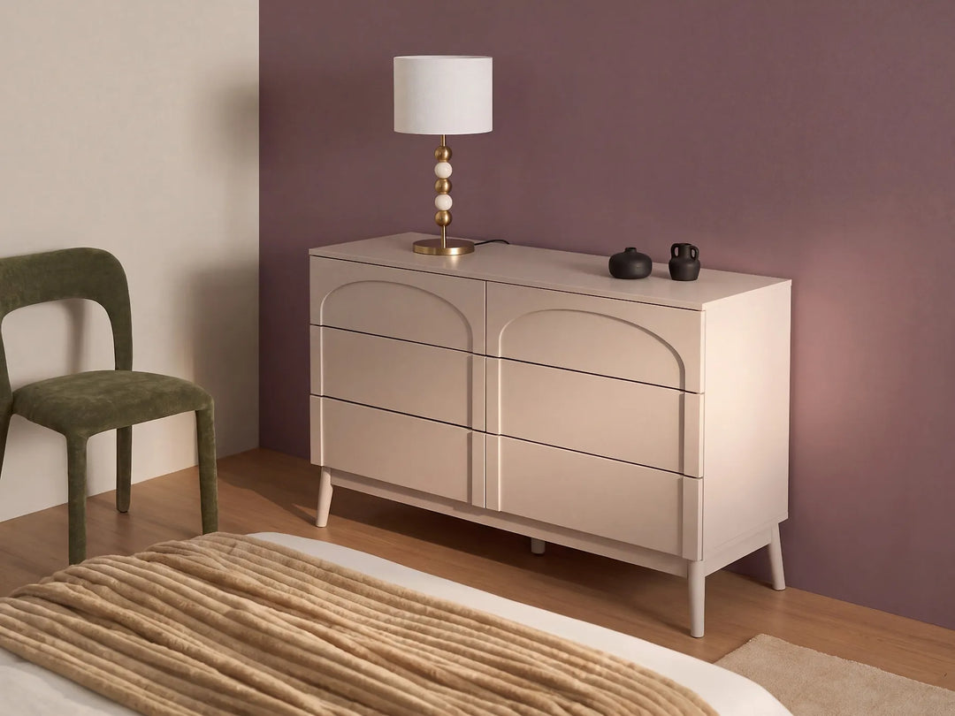 A modern six-drawer dresser in a light beige finish, styled in a bedroom setting against a mauve wall with a green chair and a bed visible.