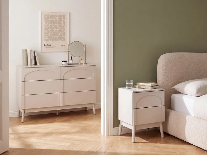 A light beige bedroom furniture set shown in a styled room, featuring a six-drawer dresser against a white wall and a matching two-drawer nightstand beside a bed with a sage green wall.