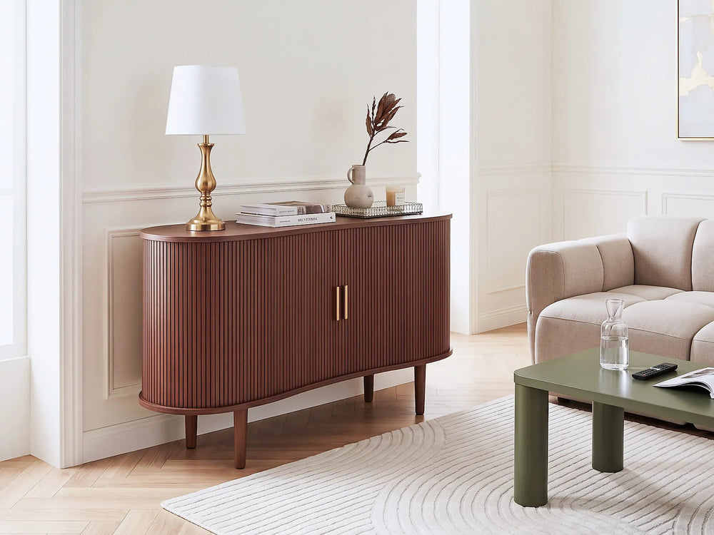 A mid-century modern sideboard cabinet in a warm reddish-brown wood finish with vertical fluting, shown in a styled living room setting next to a sofa and coffee table.