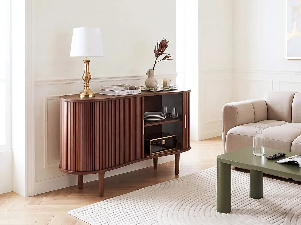 A mid-century modern sideboard cabinet in a warm reddish-brown wood finish with fluted vertical panels, shown in a bright, elegant living room next to a beige sofa and a green coffee table.