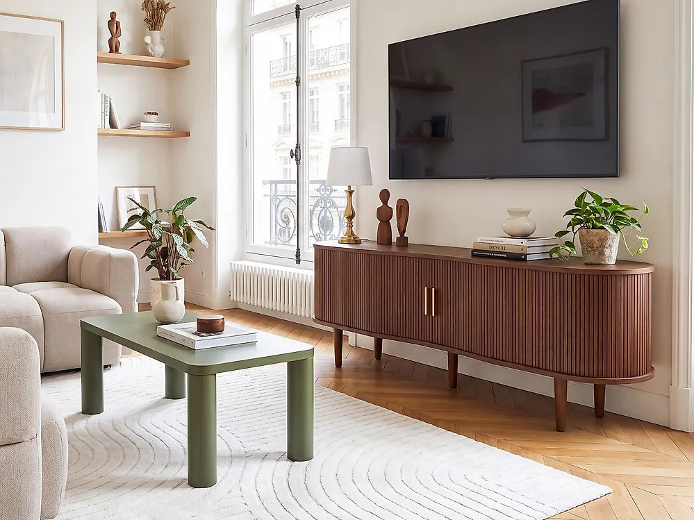 A mid-century modern curved wooden credenza or sideboard in a warm brown finish, styled in a bright living room next to a green coffee table and a beige sofa.