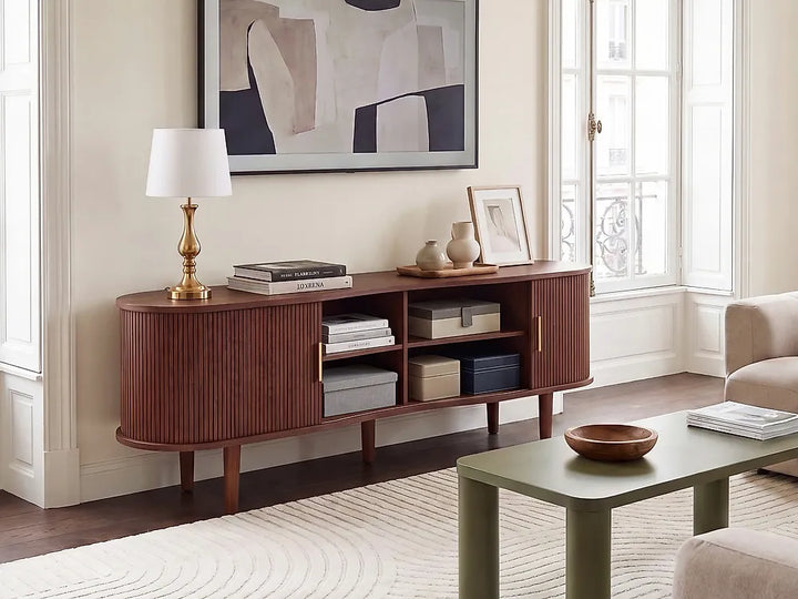 A mid-century modern wooden sideboard console in a warm reddish-brown finish, styled in a bright living room with a white table lamp, books, vases, and storage boxes.