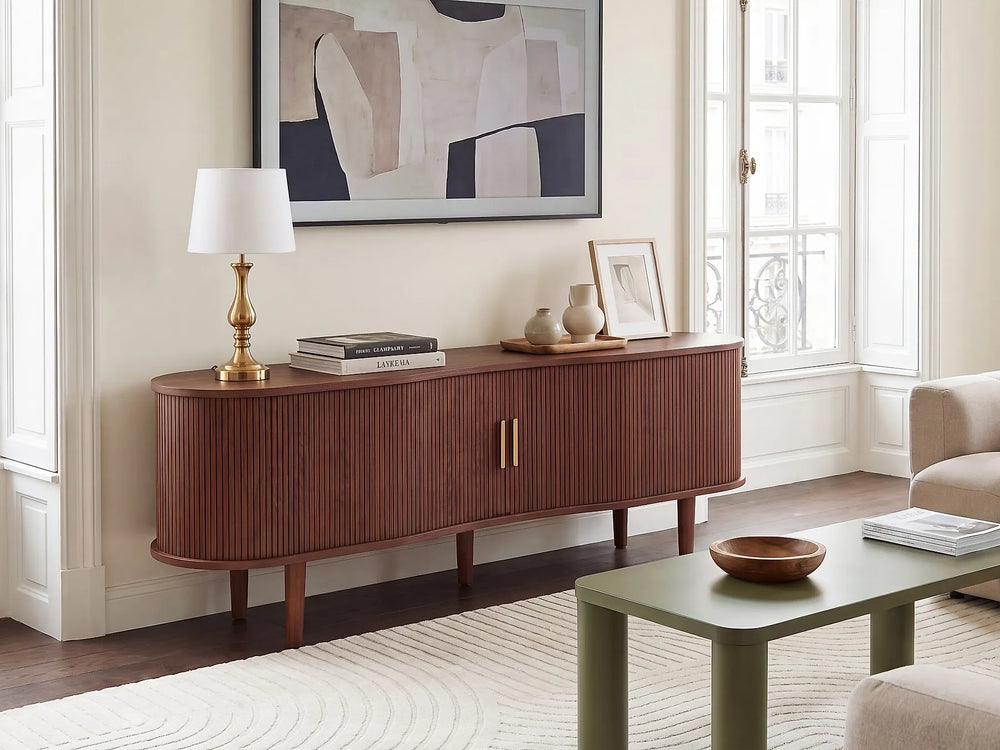 A mid-century modern wooden sideboard console cabinet in a warm reddish-brown finish, styled in a bright living room with a white table lamp, books, and decorative vases.