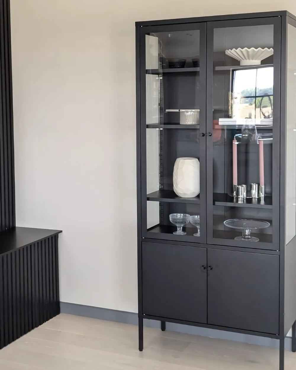 A tall black metal display cabinet with glass doors and shelves, shown in a modern room setting with light-colored walls and flooring.