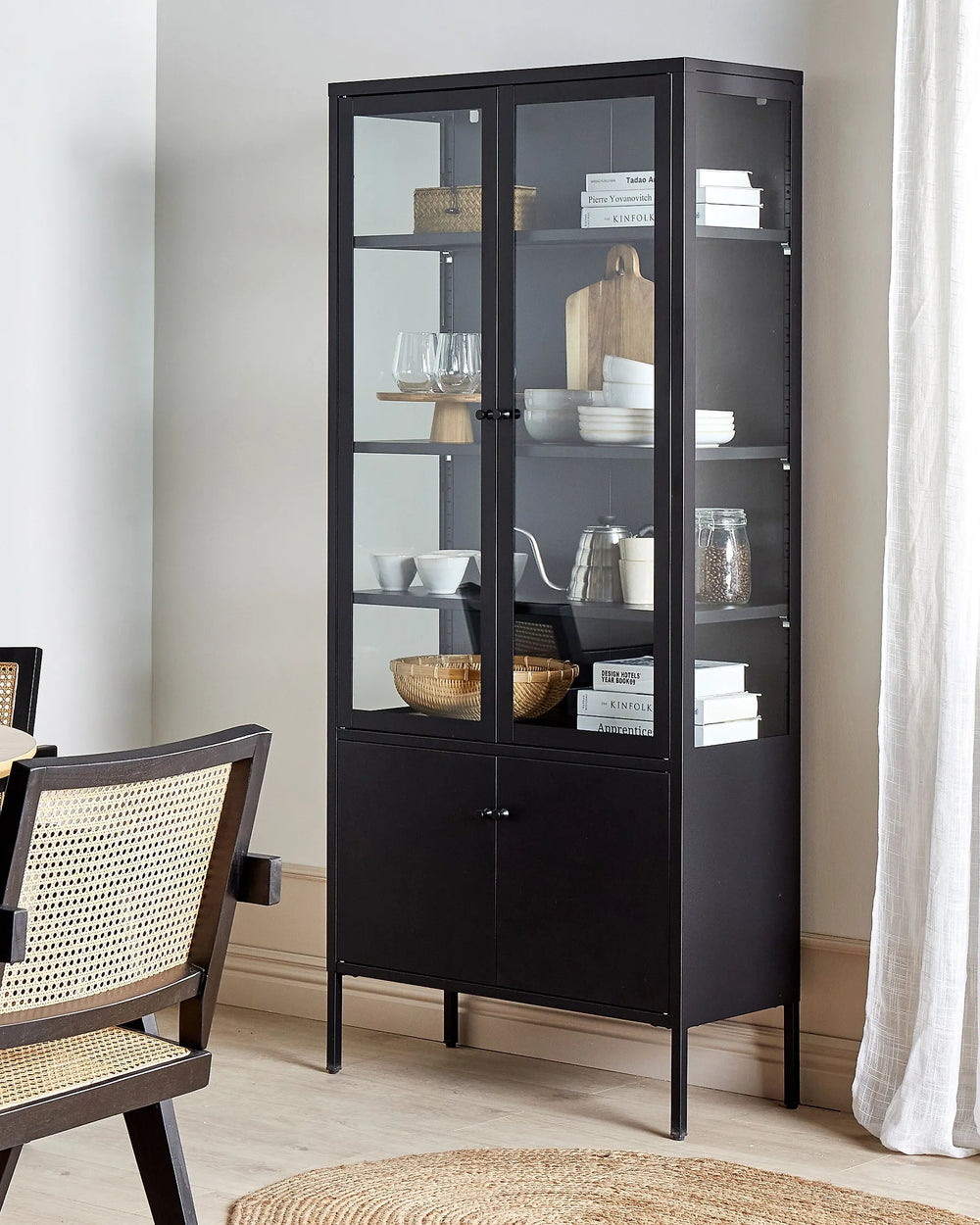 A tall black glass-front display cabinet with two lower doors, shown in a styled dining room setting next to a woven chair and curtain.