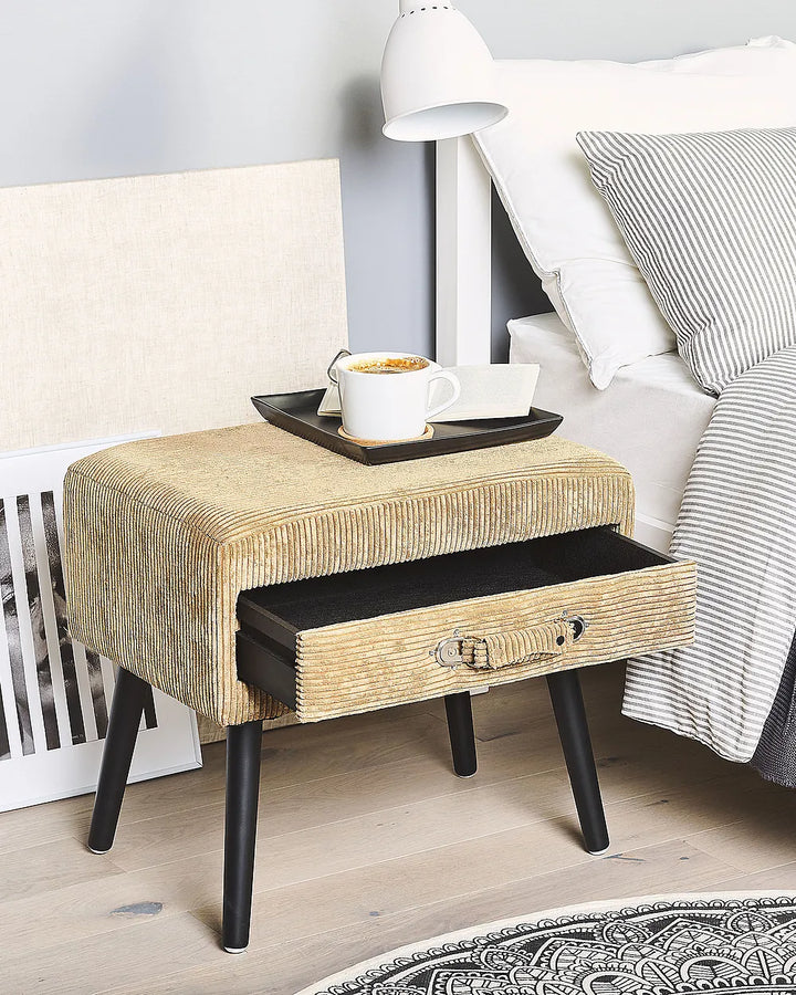 A modern upholstered bedside table with a textured beige corduroy-like fabric, black legs, and an open drawer. A coffee cup and tray sit on top, next to a bed with striped bedding in a styled bedroom setting.