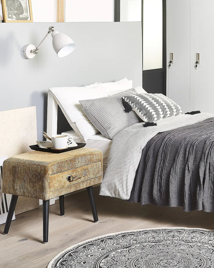 A styled bedroom scene featuring a bed with white and gray striped bedding, a textured gray throw blanket, a patterned accent pillow, and a rustic beige nightstand with black legs beside a white wall-mounted lamp.