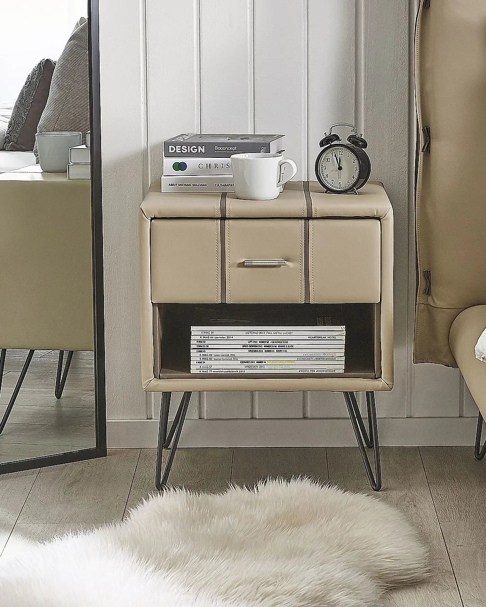 A beige upholstered bedside table with three drawers, an open lower shelf, and black hairpin legs, shown in a styled bedroom setting with books, a mug, an alarm clock, a mirror, and a white rug.