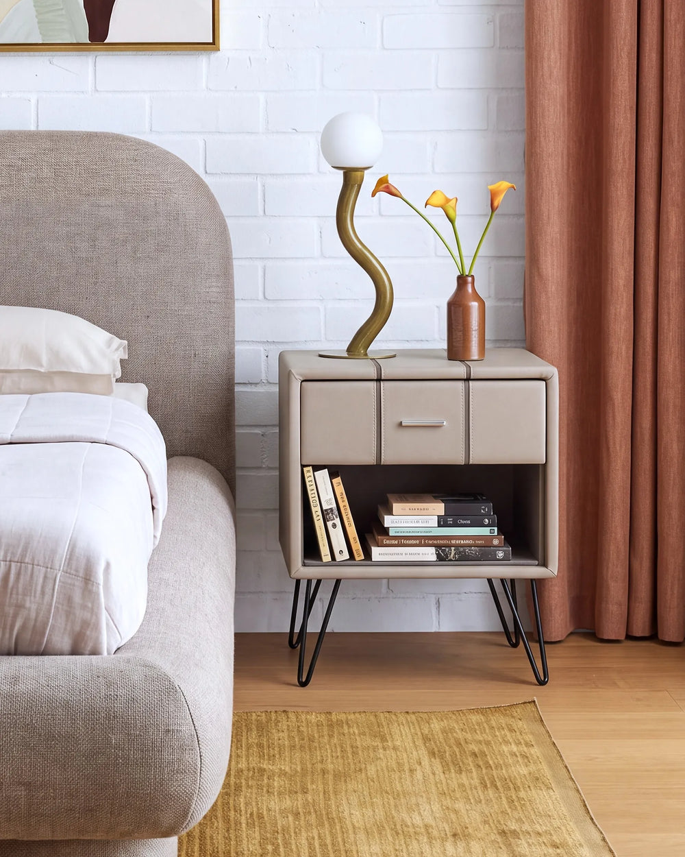 A modern bedside table with a beige upholstered front, open lower shelf, and black hairpin legs, styled in a bedroom next to a bed with white linens. The table holds a sculptural gold lamp and a small vase with yellow flowers.