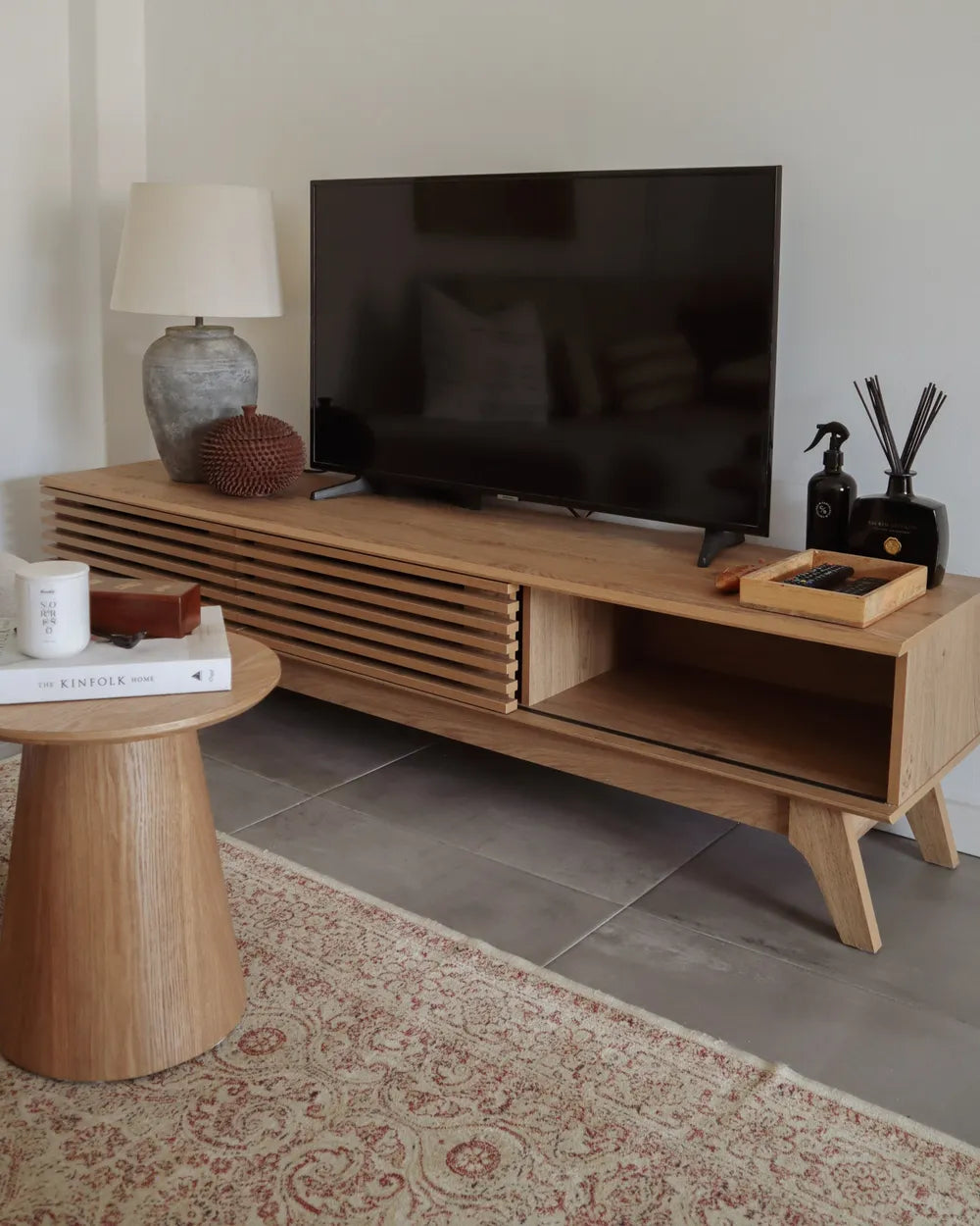 A light oak wood TV console with a slatted front and open storage shelves, styled in a living room setting with a flat-screen TV, a side table, and a rug.
