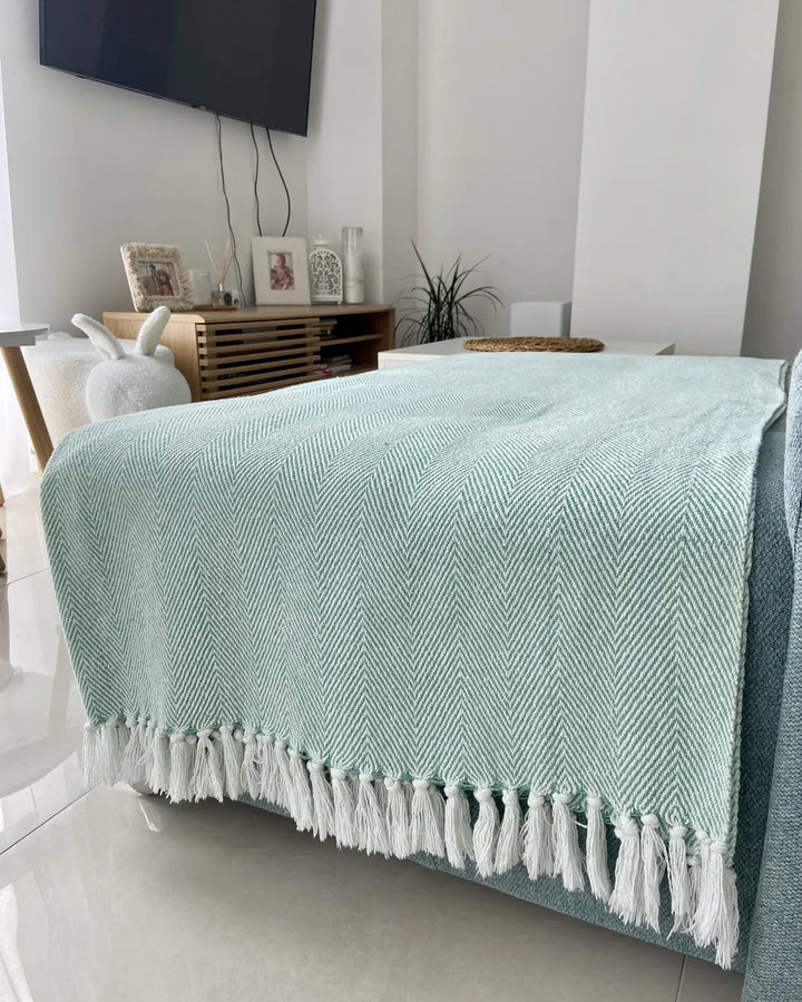 A mint green and white herringbone patterned throw blanket with white tassels, draped over a bed in a modern bedroom setting.