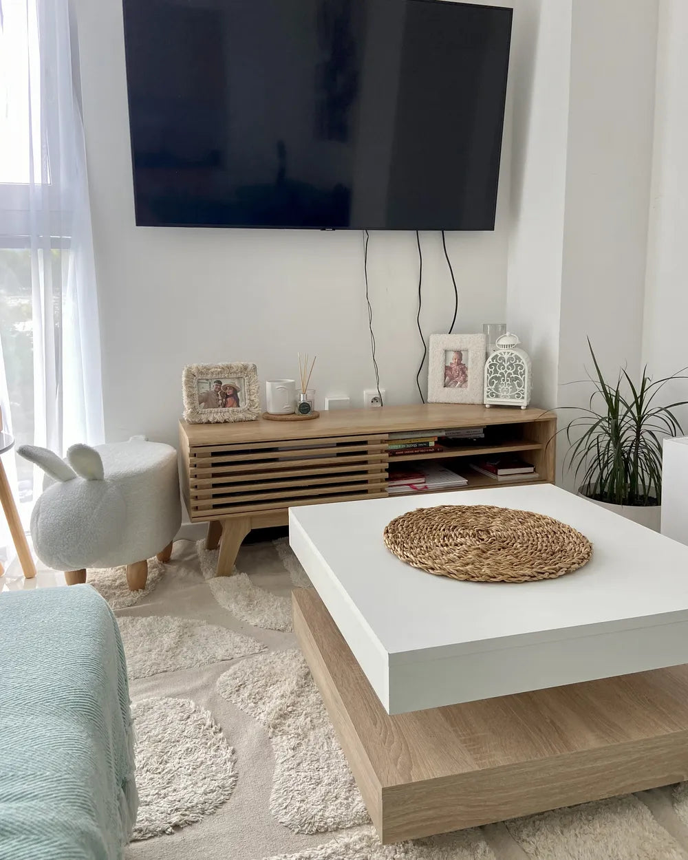 A modern living room setup featuring a white and light wood coffee table in the foreground, a light wood media console with a black wall-mounted TV behind it, and a white bunny-shaped stool on a patterned cream rug.