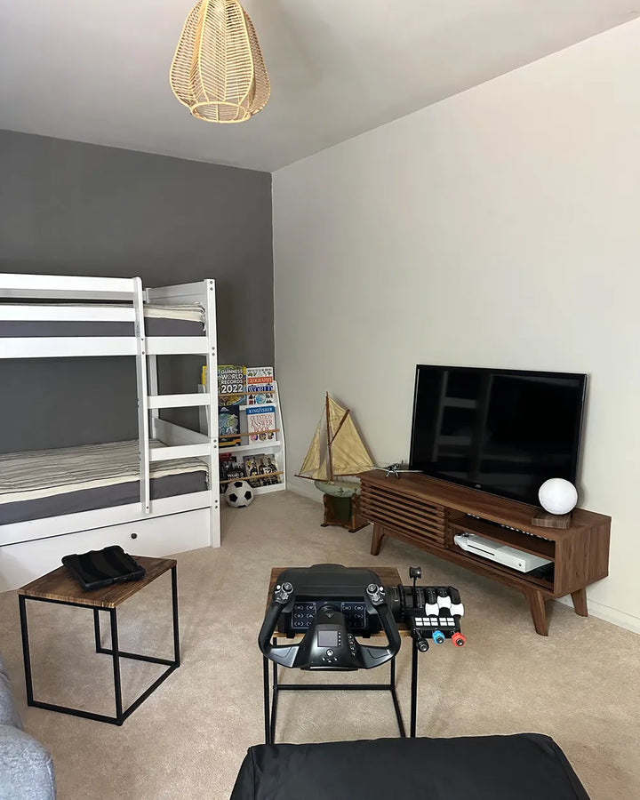 A child's bedroom or playroom featuring a white wooden bunk bed, a dark gray accent wall, a beige wall, a wooden TV stand with a flat-screen television, a small side table, and a central glass coffee table.