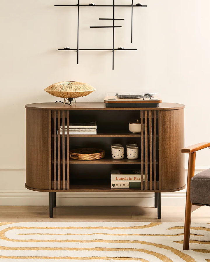 A mid-century modern wooden sideboard cabinet with open shelving and vertical slat details, styled with home decor items in a room setting with a patterned rug and chair partially visible.