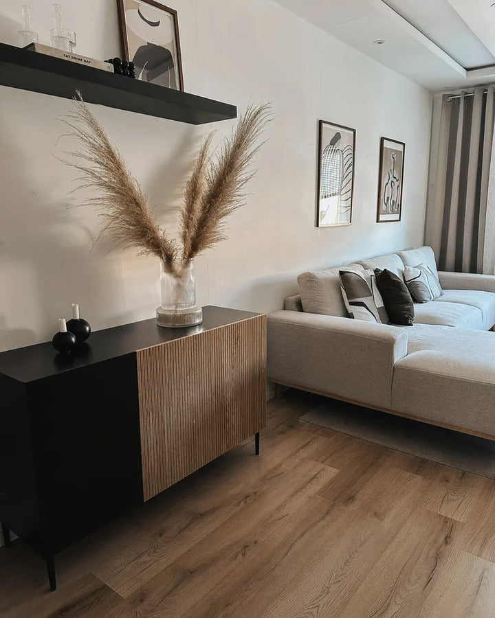 A modern living room featuring a black and light wood sideboard cabinet with a vase of pampas grass, next to a light gray sectional sofa with neutral-toned pillows.