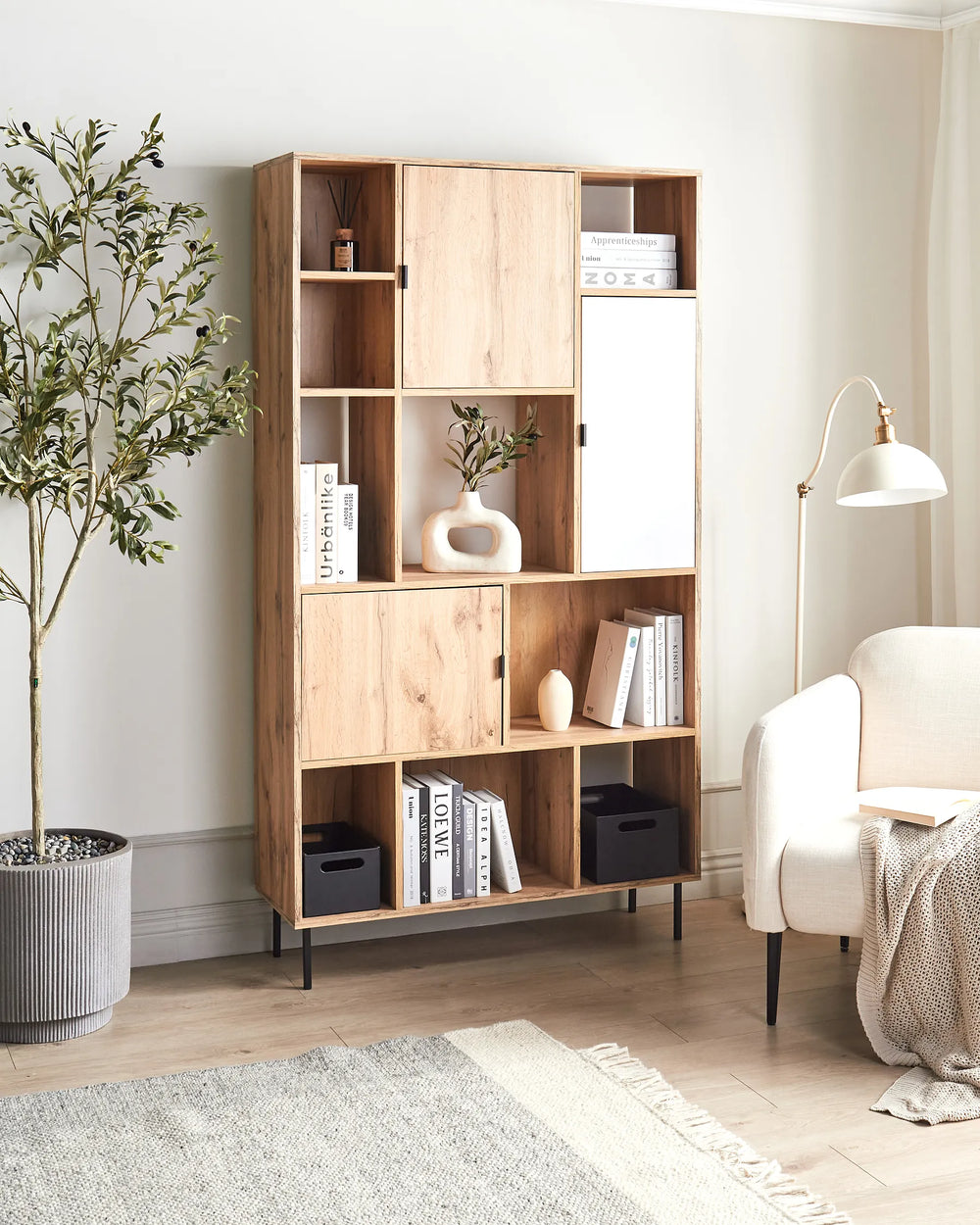 A tall, modern wooden bookcase with an open grid design, styled with books, vases, and plants in a bright, contemporary living room next to a white armchair and a floor lamp.