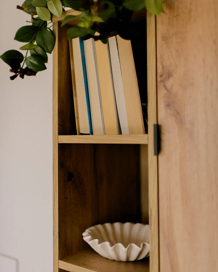 A close-up of a light natural wood cabinet or shelf unit with a door, holding several books on the top shelf and a white scalloped bowl on the bottom shelf, with green plant leaves hanging over the top.