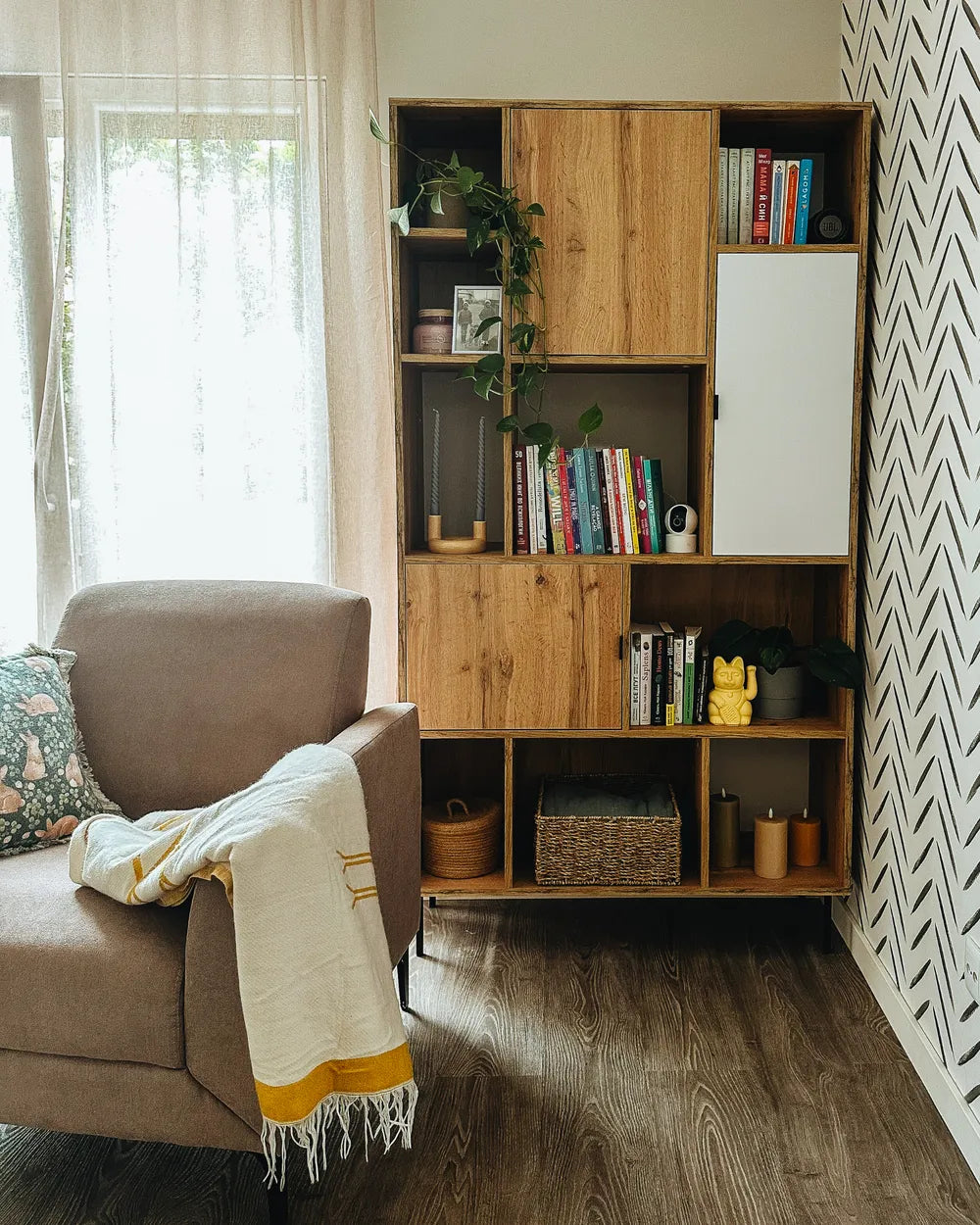 A tall wooden bookcase with mixed wood and white cabinet doors, styled with books, plants, and baskets in a bright living room next to a brown armchair.