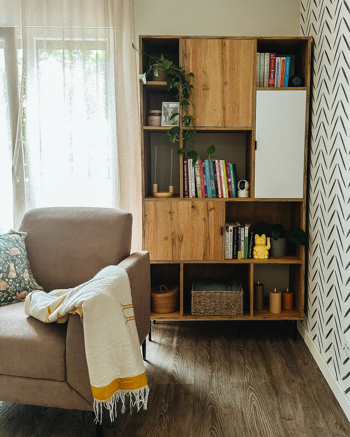 A tall wooden bookcase with mixed wood and white cabinet doors, styled with books, plants, and baskets in a bright living room next to a brown armchair.