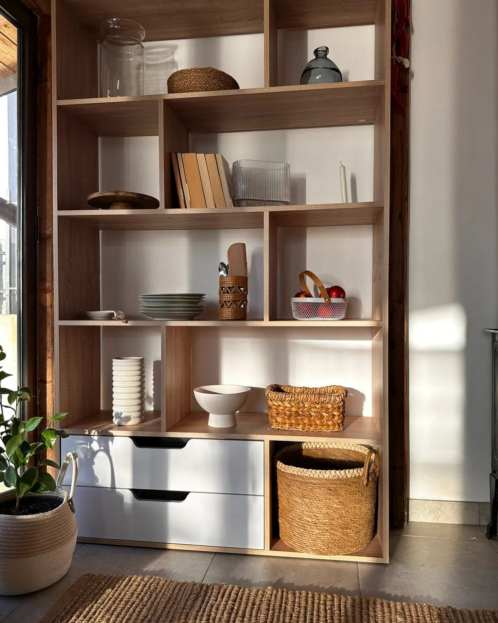 A tall wooden open shelving unit with multiple compartments, styled with books, bowls, baskets, and storage bins, set in a sunlit room with a potted plant nearby.