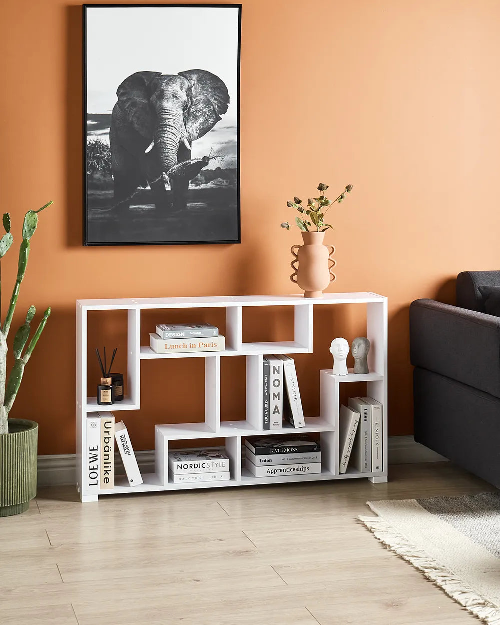 A white modular open bookcase styled in a living room setting against a warm terracotta wall, holding books, a small vase, and decor.