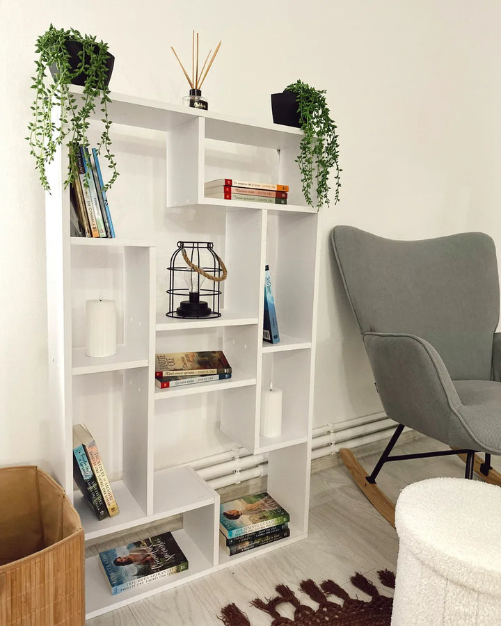 A white modular open bookshelf styled with books, potted trailing green plants, candles, and a small black metal birdcage, positioned next to a gray armchair in a bright room.