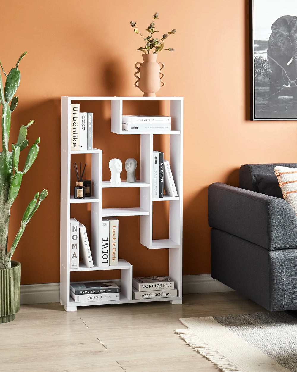 White bookshelf against an orange wall with books and decor items, next to a gray sofa and potted plant.