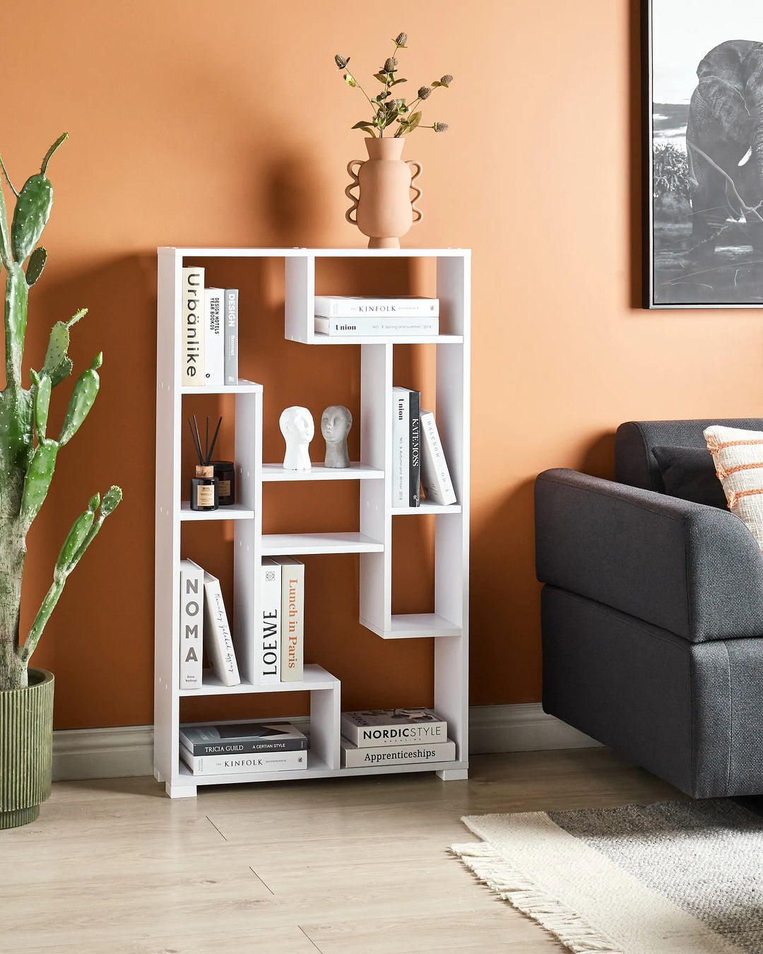 White bookshelf against an orange wall with books and decor items, next to a gray sofa and potted plant.